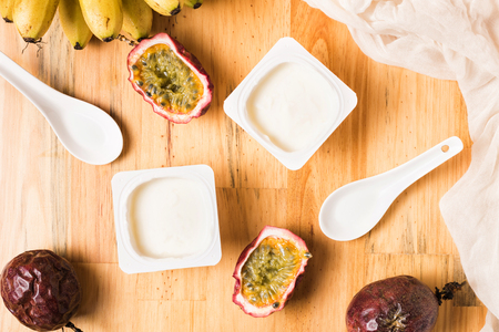 Plastic cups with yogurt on wooden background. Breakfast with bananas, passion fruits and yoghurt. White spoon and napkin on table. Top viewの写真素材