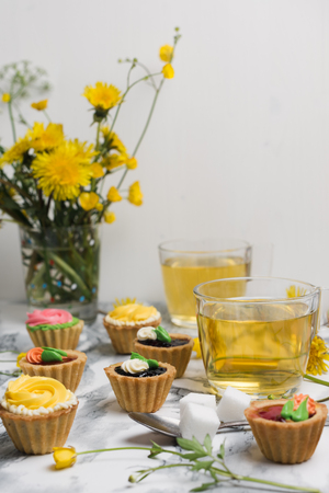 Colorful cupcakes on a gray marble table. Cakes with cream and jam. Cup of green tea and yellow flowers on a backgroundの写真素材