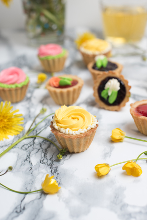 Colorful cupcakes on a gray marble table. Cakes with cream and jam. Cup of green tea and yellow flowers on a backgroundの写真素材