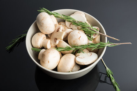 Common mushroom in a bowl. Sliced ??champignon and rosemary in a white plate on a black backgroundの写真素材