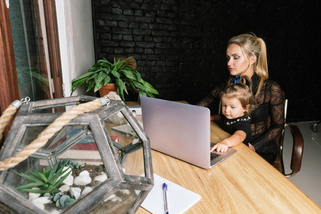 Beautiful business mother with her little girl. Young blonde woman in classic suit is smiling while playing with her cute baby in home office.の写真素材