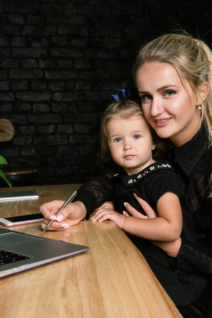 Beautiful business mother with her little girl. Young blonde woman in classic suit is smiling while playing with her cute baby in home office.の写真素材