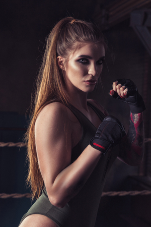 Young pretty boxer woman standing on ring. Female fighter in black glove training in dark gymの写真素材