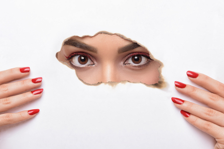 Eyes looking through the hole of the wall. Woman with fashion make up and red manicure Peeking in a slot in a burned sheet of white paperの写真素材