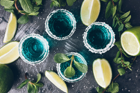 Alcohol shots on an old metal tray. Shooter glasses of blue vodka or Tequila on black table. Mojito with mintの写真素材
