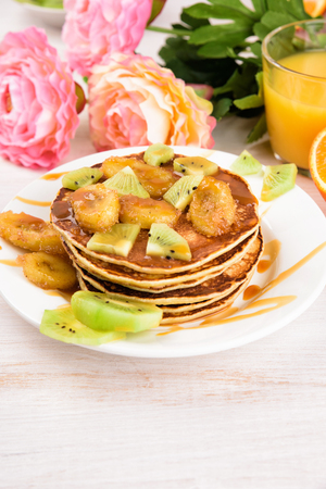 Stack of pancakes with a grilled bananas and a caramel. Glass of juice, orange, kiwi slices. Pink flowers, flat lay, top viewの写真素材