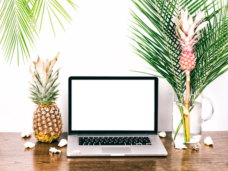 Laptop with a blank white screen. Freelancer desktop whith computer, palm branches and decorative pink pineapple in a vase on a wooden table.の写真素材