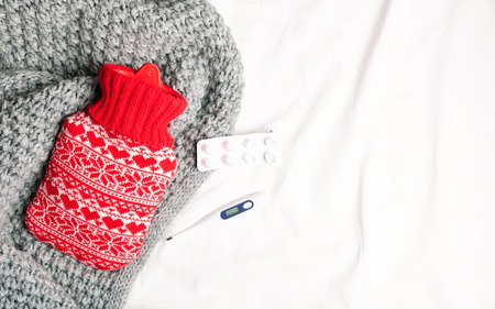 Red hot-water bottles and thermometer, drugs on a bed. Heat pads on sheet, warm cozy grey knitted...