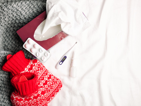 Red hot-water bottles and thermometer, drugs on a bed. Heat pads on sheet, warm cozy grey knitted plaid and box of papper handkerchief. Top view or flat lay, copy space for your textの写真素材