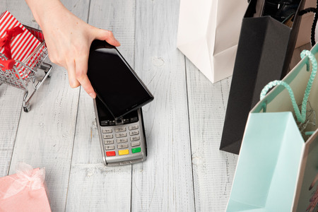 Woman pays with NFC technology on a smartphone. Payment terminal on white wooden background. Laptop...