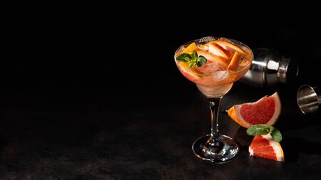Grapefruit cocktail in a martini glass with mint and ice cubes. Cold summer citrus fresh beverage and shaker on dark backgroundの写真素材