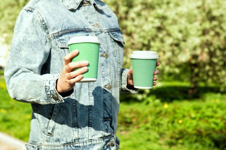 Female hands holding coffee mint color paper cups. Woman in light blue jeans jacket holds a drink on the street. Summer sun, green grassの写真素材