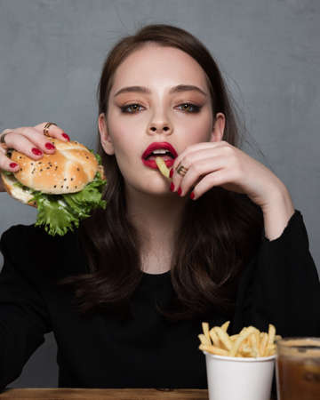 Glamour woman with a perfect fashion red manicure and makeup eating burger and French fries.