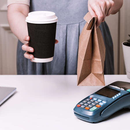 Woman holding ordering in a cafe. NFC payment terminal and take away cup of coffee with on a white table. Credit card or phone pay pos banking device, card machine.の写真素材