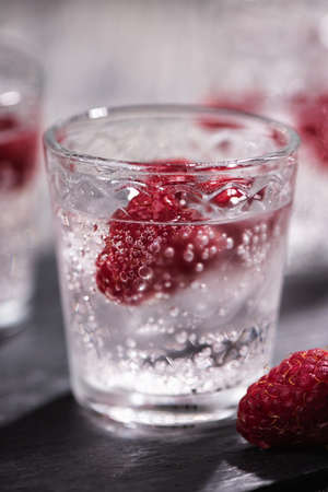 Alcohol shots of berries cocktail with a raspberry on black stone tray. Glass of sparkling water on...