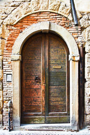 Old wooden door with a metal knocker and letter slot - wonderful texture. The door has little metal studs. On the side, there is a modern door bell and intercom. Exposed brick and pipes and a stone arch complete the picture.の写真素材
