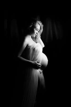 Pregnant woman in the studio on a black background, standing half-turn knee-deep. In a white shirt and underwear, hands on a bare stomach.Bare shoulders.One knee bent. Head tilted, looks at the floorの写真素材