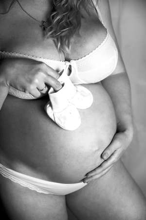 Pregnant woman in the studio on a black background, standing half-turn knee-deep. In a white shirt and underwear, hands on a bare stomach.Bare shoulders.One knee bent. Head tilted, looks at the floorの写真素材