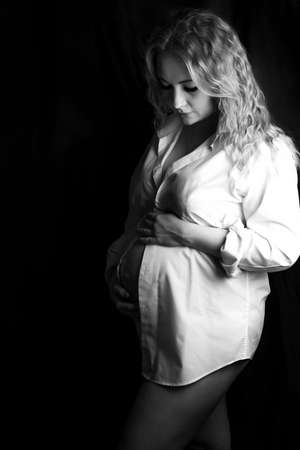 Pregnant woman in the studio on a black background, standing half-turn knee-deep. In a white shirt and underwear, hands on a bare stomach.Bare shoulders.One knee bent. Head tilted, looks at the floorの写真素材