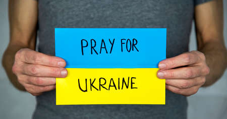 A standing man on a gray, holds in his hands background a yellow-blue cardboard Ukrainian flag with the inscription "Pray for Ukraine"の写真素材