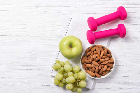Fitness and healthy eating concept, pink dumbbells with apple, grapes and almond nuts on notepad, wooden background, top view with copy spaceの写真素材