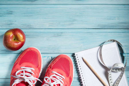 Fitness concept, pink sneakers, red apple and notebook with pencil and measuring tape on wooden background, top viewの写真素材