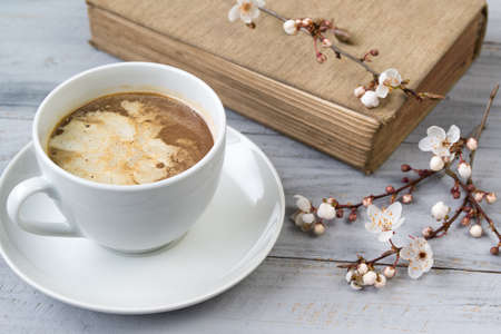 Cup of coffee with cream and the old book with blooming cherry branches on wooden backgroundの写真素材