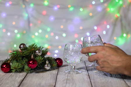 Hand holding a glass of champagne. Two glasses of champagne and red Christmas-tree decorations on a table again christmas fairy lights bokeh on background.の写真素材