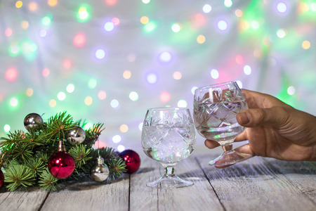 Hand holding a glass of champagne. Two glasses of champagne and red Christmas-tree decorations on a table again christmas fairy lights bokeh on background.の写真素材