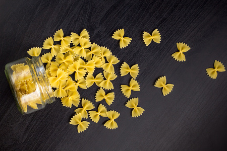 Glass jar filled with dry farfalle pasta and scattered around on dark black backgroundの写真素材