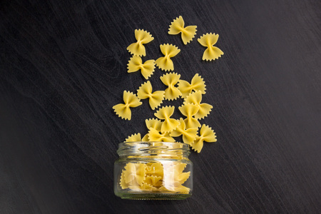 Glass jar filled with dry farfalle pasta and scattered around on dark black background