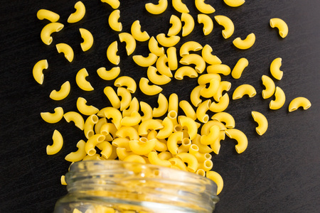 Glass jar filled with dry pasta horns and scattered around on dark black backgroundの写真素材