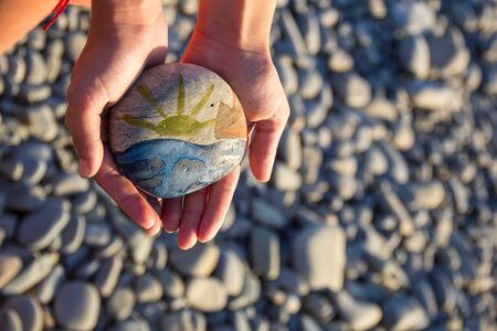 Bright sun painted on pebble in the hands of a child on the background of a pebble beach in summerの写真素材