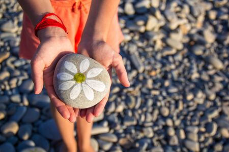 pebbles with a painted daisy in the hands of a child on the background of a pebble beach in summer