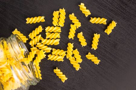 Glass jar filled with dry pasta spiral fusilli and scattered around on dark black background