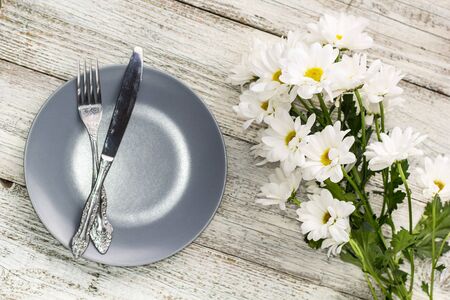 empty plate with fork and knife decorated with chamomile flowers on white wooden background. Top...