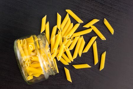 Glass jar filled with dry penne pasta and scattered around on dark black backgroundの写真素材