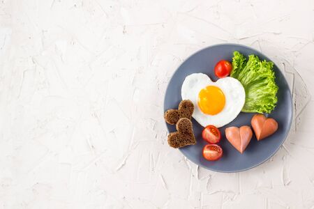 Fried eggs in the shape of a heart with sausages, herbs and tomatoes. Breakfast for your loved ones on Valentine's day on white background. Top view with copy spaceの写真素材