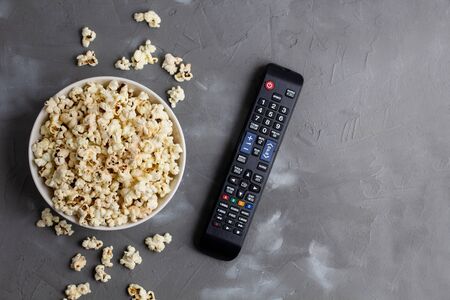 A white bowl of popcorn and TV remote on a grey background. The concept of watching TV, film, TV series, sports, shows.