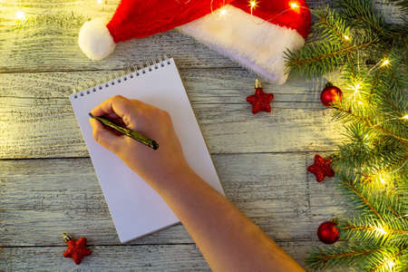 hand of woman writing letter on notepad paper with Christmas decorations and bokeh on white wooden backgroundの写真素材