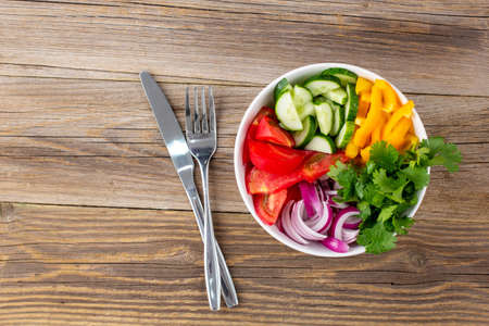 Plate of rainbow salad with different vegetables and herbs in white bowl with knife and fork on natural wooden background Top view with copyspaceの写真素材