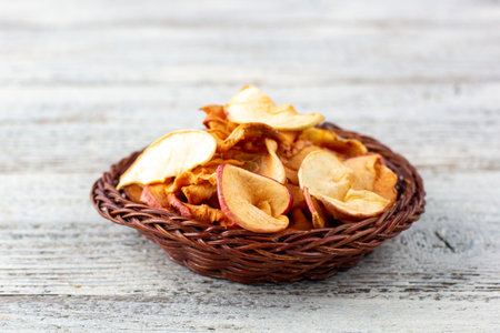 A pile of dried slices of apples in wicker basket on white wooden background. Dried fruit chips. Healthy vegan foodの写真素材