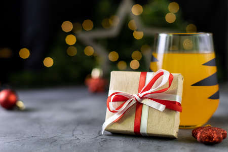 Christmas gift in craft paper with white and red ribbon and orange cocktail in glass with black stripes on dark background with star and golden bokehの写真素材