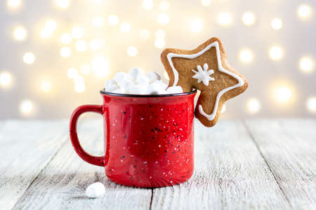 A red cup of winter hot drink with marshmallows and gingerbread star on a white wooden background. Traditional Christmas atmosphere with bokeh. New year holiday concept. Festive backgroundの写真素材