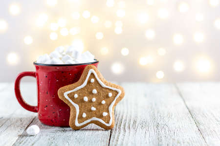 A red cup of winter hot drink with marshmallows and gingerbread star on a white wooden background. Traditional Christmas atmosphere with bokeh. New year holiday concept with copy spaceの写真素材