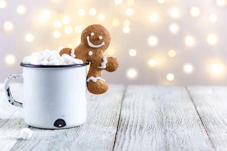 A white cup of winter hot drink with marshmallows and gingerbread man on a white wooden background. Festive xmas background. Traditional Christmas atmosphere with bokeh. New year holiday concept.の写真素材