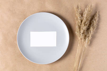 Blank white paper card on plate for mock up with dried spikelets decoration on craft beige background. Flatlay, top view with copy space, mockup. Minimal boho styleの写真素材