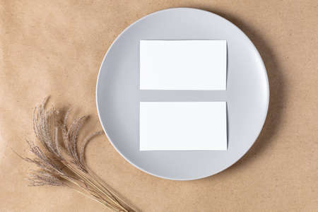 Blank white paper card on plate for mock up with dried spikelets decoration on craft beige background. Flatlay, top view with copy space, mockup. Minimal boho styleの写真素材