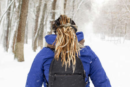 Back view of a woman with dreadlocks hairstyle. Woman with black backpack and blue jacket on winter backgroundの写真素材