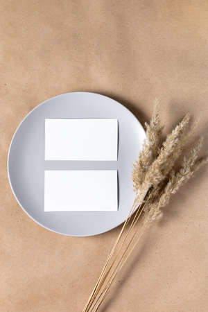 Blank white paper card on plate for mock up with dried spikelets decoration on craft beige...